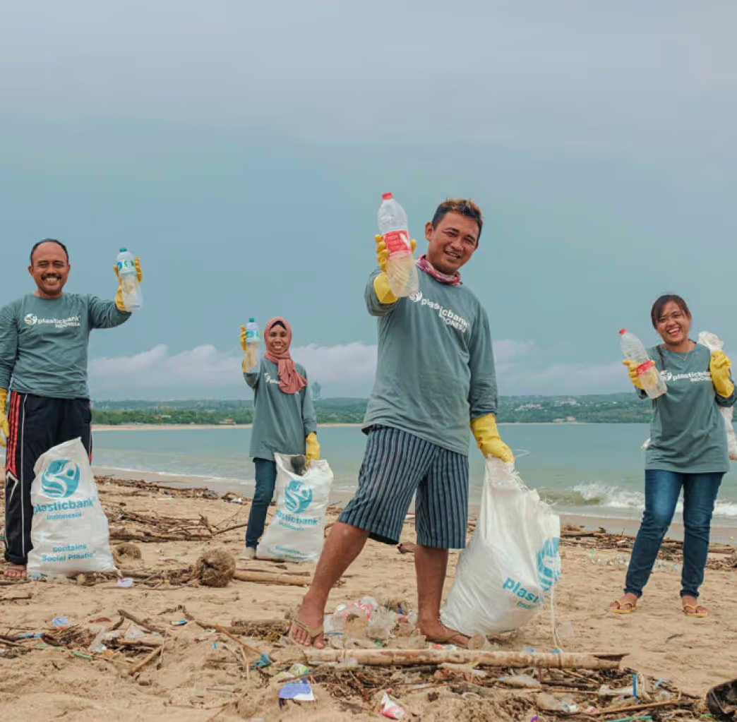 Four volunteers in Plastic Bank shirts and yellow gloves hold plastic bottles and bags while cleaning a beach.