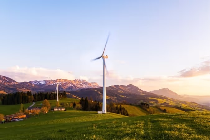 Wind turbines on green hills with mountains in the background at sunset.